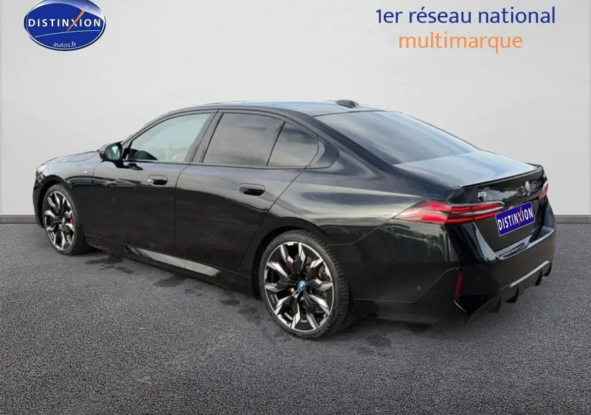 BMW Série 5 i5 M60 noire vue 3/4 arrière droit, avec jantes alu bicolores et feux arrière LED allumés.