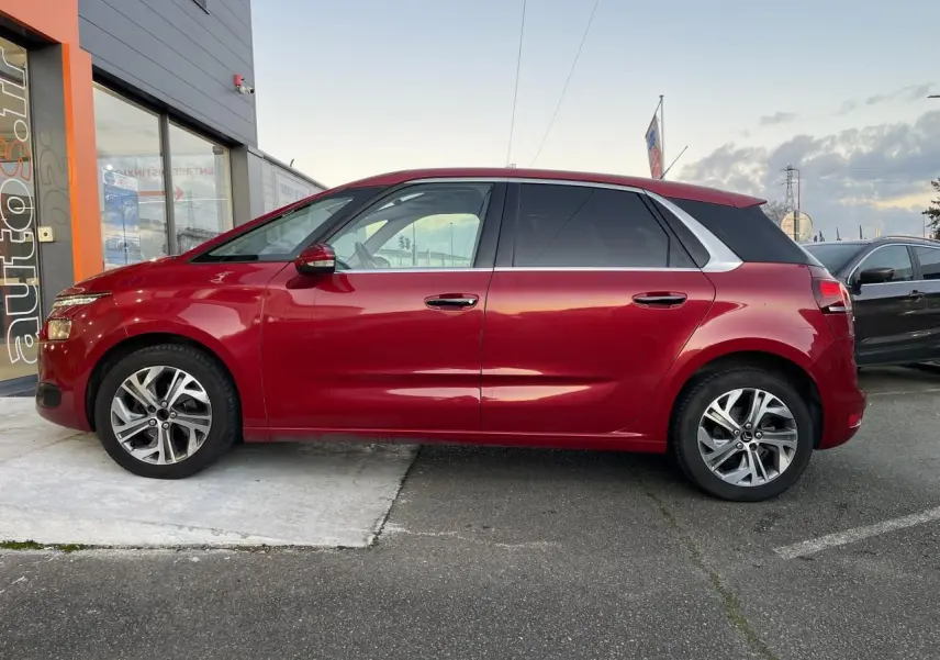 Profil côté gauche d'une Citroën C4 Picasso rouge foncé garée devant un bâtiment commercial.