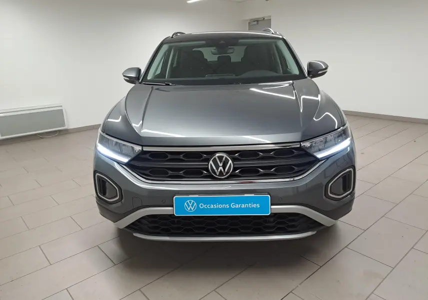 Vue frontale d'un Volkswagen T-Roc gris Indium avec feux LED allumés et calandre noire dans un intérieur neutre.