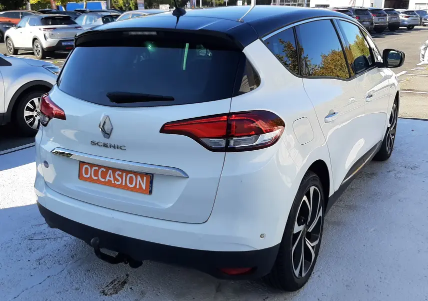 Vue 3/4 arrière droite d'un Renault Scénic IV blanc nacré avec toit noir, en stationnement extérieur sur un parking.