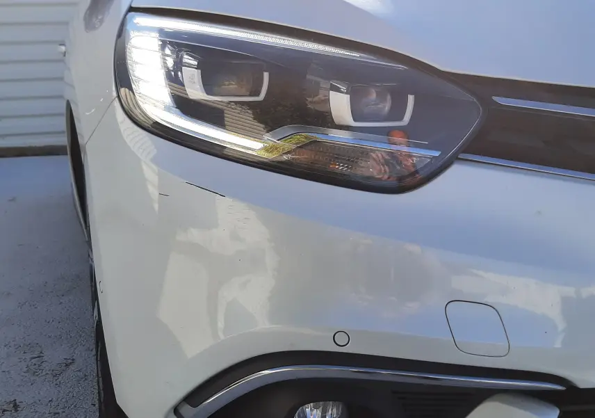 Gros plan sur le phare avant gauche d'un Renault Scénic IV blanc nacré avec toit noir, mettant en valeur ses feux LED et détails chromés.