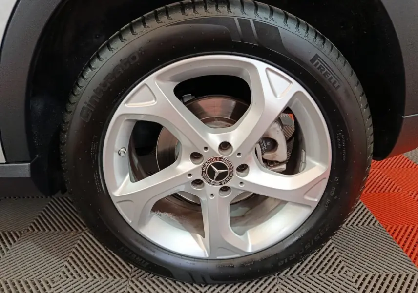 Gros plan sur la roue arrière droite d'un Mercedes GLA 200 d gris clair, avec partie de la carrosserie visible sur fond de sol texturé.