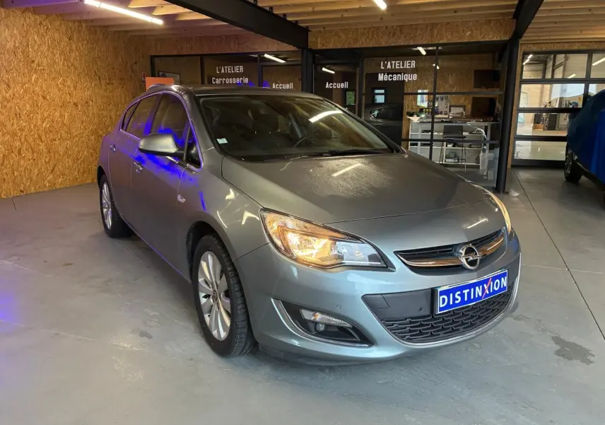 Opel Astra gris 2015 vue 3/4 avant droit avec phares allumés dans un atelier mécanique.