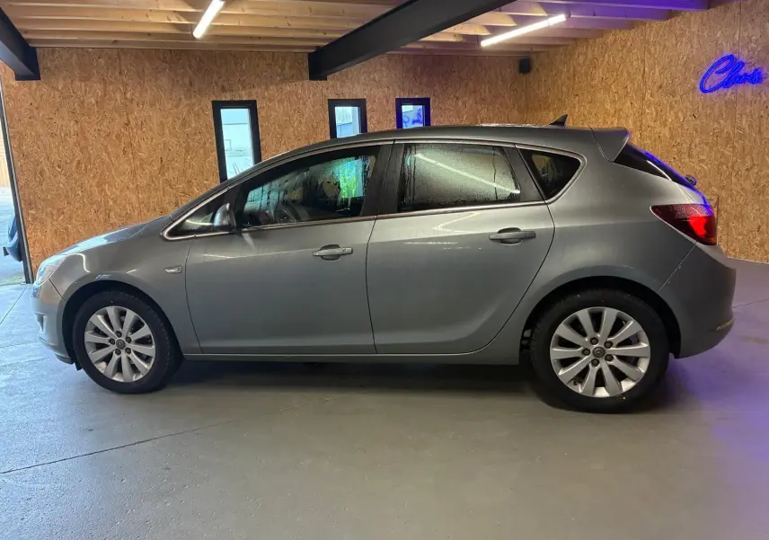 Vue latérale droite d'une Opel Astra gris métallisé de 2015, avec jantes alliage et vitres légèrement embuées.