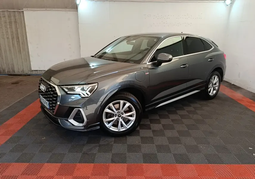 Audi Q3 Sportback gris clair en 3/4 avant droit, avec jantes alliage et prise de recharge visible côté avant gauche.