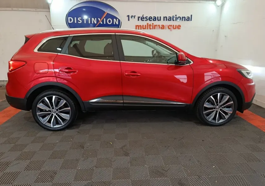 Vue latérale droite d'un Renault Kadjar Intens rouge brillant, avec jantes alliage et toit chromé en arrière-plan showroom.