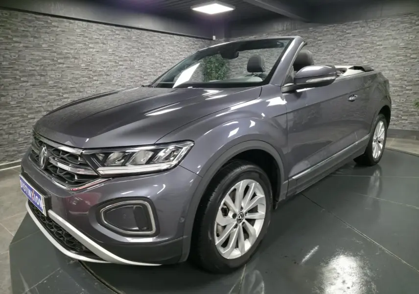 Volkswagen T-Roc Cabriolet gris métallisé en 3/4 avant droit, capote noire ouverte et jantes alliage visibles.