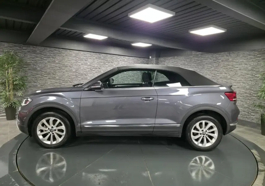 Volkswagen T-Roc Cabriolet gris métallisé vue de profil côté gauche avec capote noire fermée en intérieur showroom.