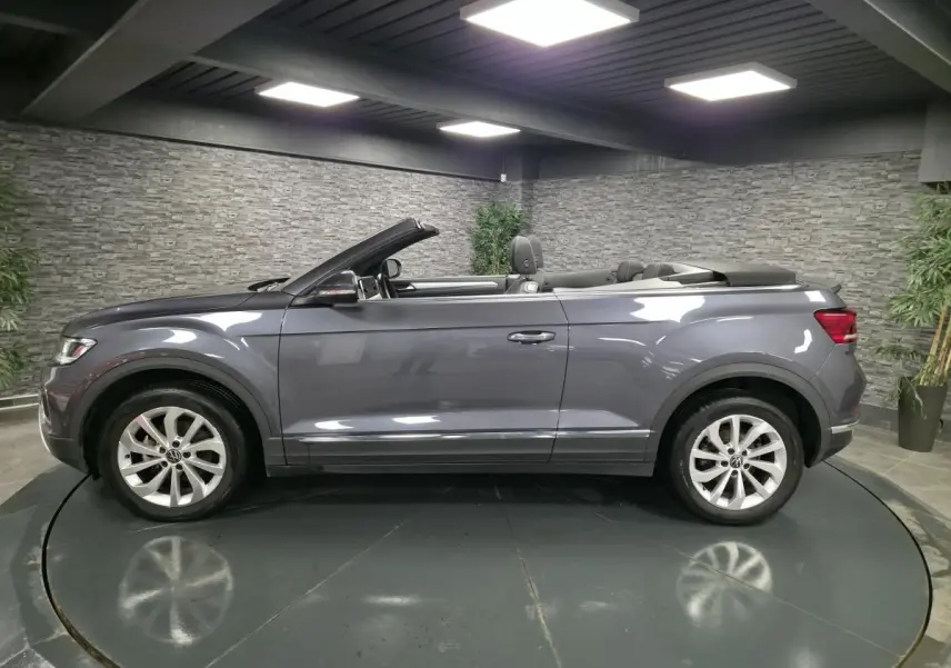 Profil côté gauche d'un Volkswagen T-Roc Cabriolet gris métallisé 2024 avec capote ouverte en intérieur showroom.