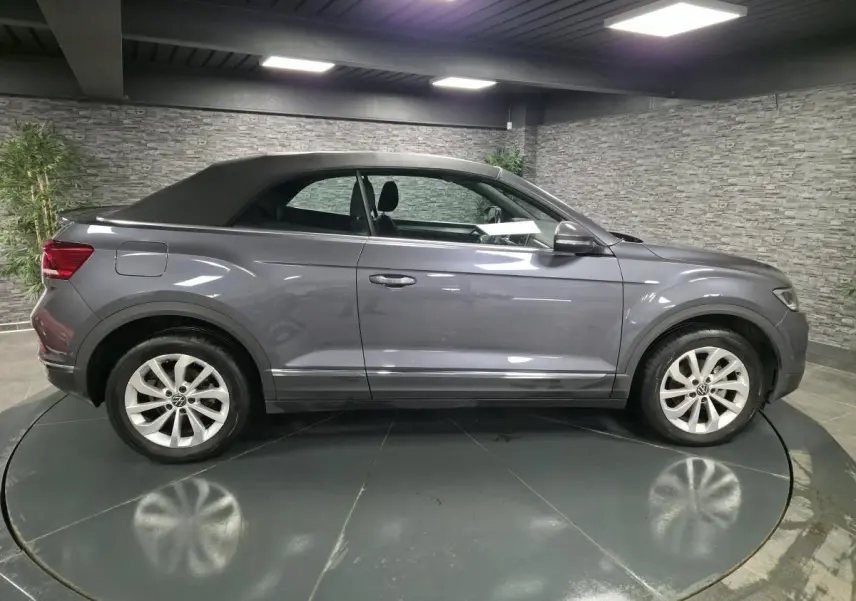 Profil côté gauche d'un Volkswagen T-Roc Cabriolet gris métallisé avec capote noire dans un showroom.