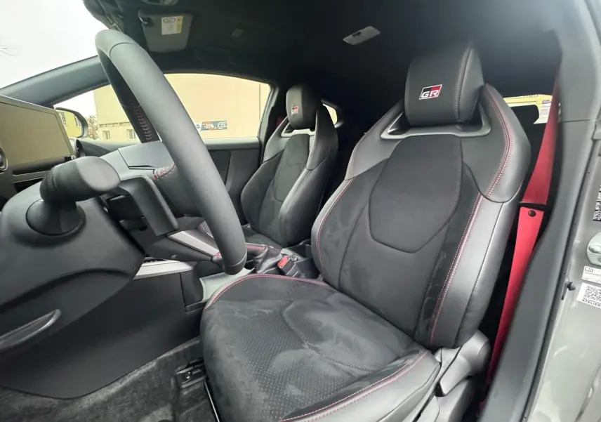 Vue intérieure côté conducteur de la Toyota Yaris GR avec sièges sport noirs, surpiqûres rouges et ceinture rouge visible.