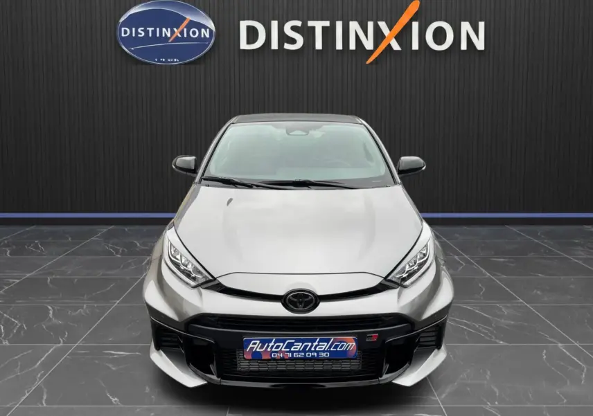 Vue avant d'une Toyota Yaris GR gris platine avec calandre noire et logo GR, sur fond noir avec logo Distinxion.