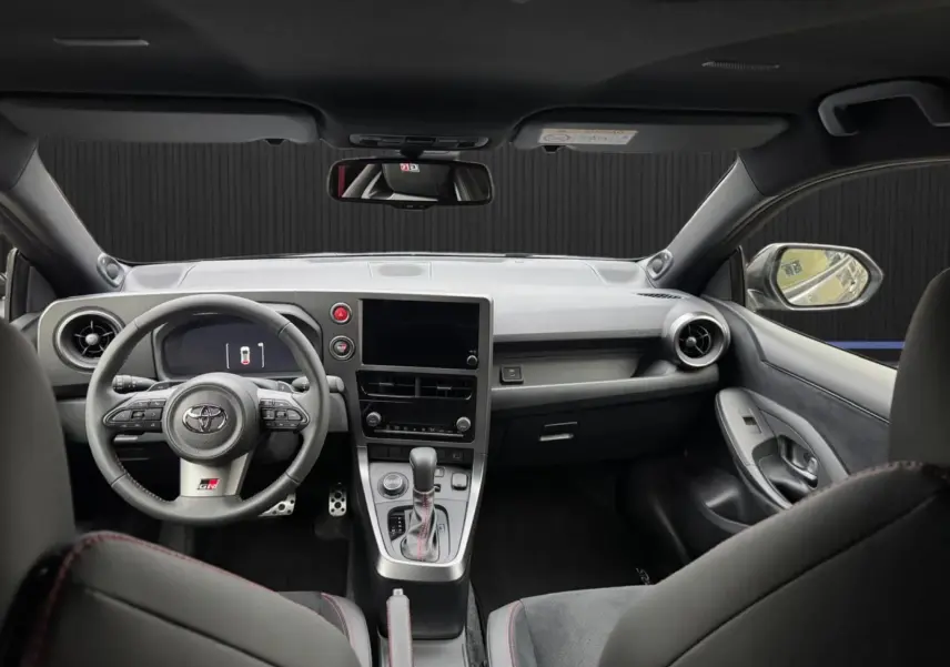 Vue intérieure avant de la Toyota Yaris GR Pack Techno, tableau de bord noir avec surpiqûres rouges et volant sport multifonction.
