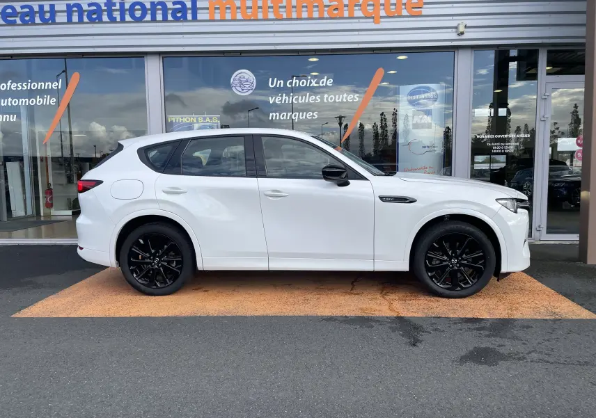 Vue de profil côté gauche d'un Mazda CX-60 blanc nacré avec jantes noires, stationné devant un showroom multimarque.