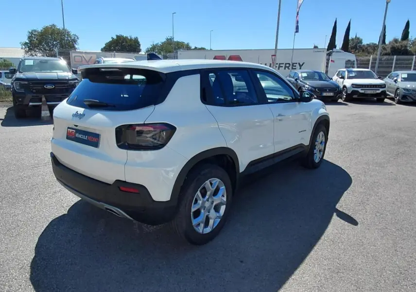 Jeep Avenger blanc vu en 3/4 arrière droit sur un parking, avec des jantes alliage et des feux arrière fumés.