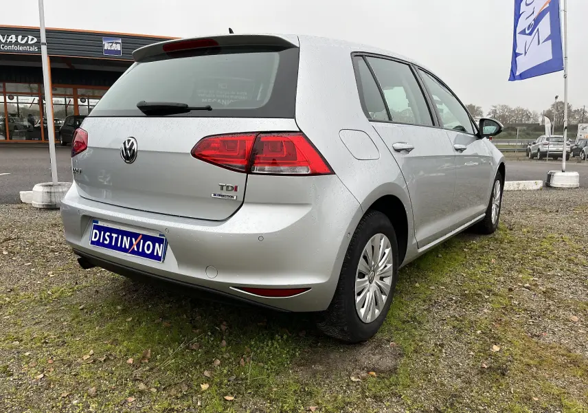 Vue 3/4 arrière droite d'une Volkswagen Golf 7 argent avec badge TDI et plaque Distinxion sur un parking extérieur.