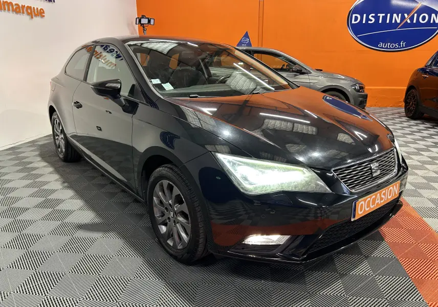 SEAT Leon noir vue 3/4 avant droit, phares allumés, sur sol damier dans showroom avec panneau orange "Occasion".