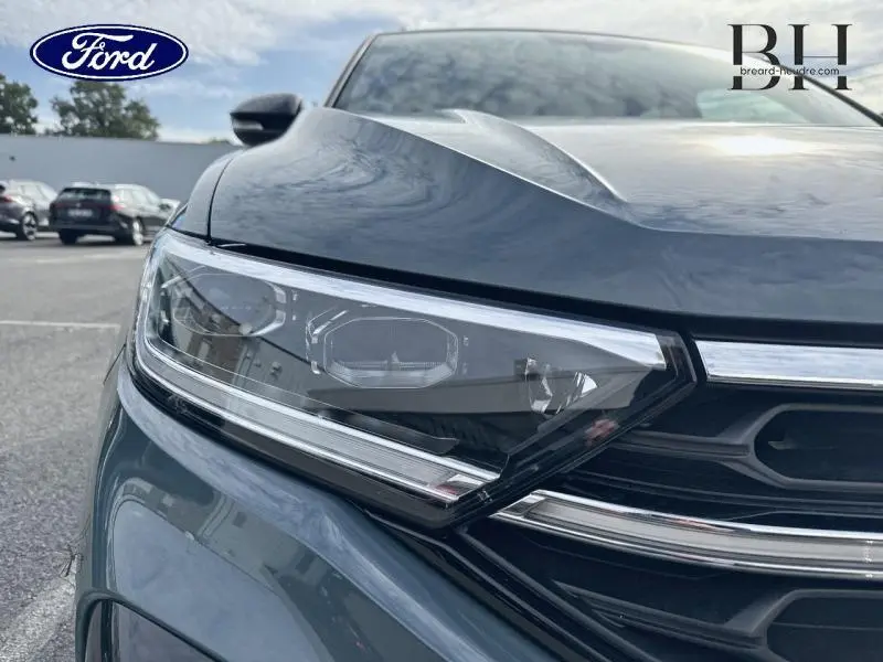 Gros plan sur le phare avant gauche d'un Volkswagen T-Roc bleu pétrole métallisé, soulignant les détails du bloc optique et la calandre.