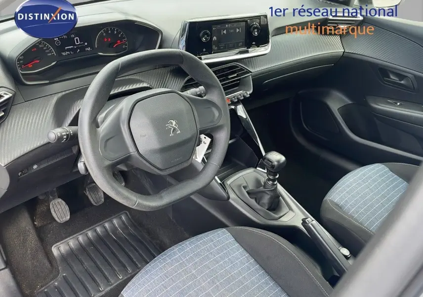 Vue intérieure côté conducteur du Peugeot 208 blanc banquise 2021, volant compact et sièges tissu bleu noir rayé.