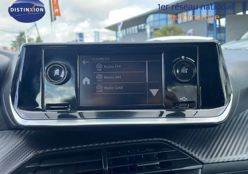 Écran tactile central du tableau de bord de la Peugeot 208 blanche, avec commandes audio et climatisation visibles.