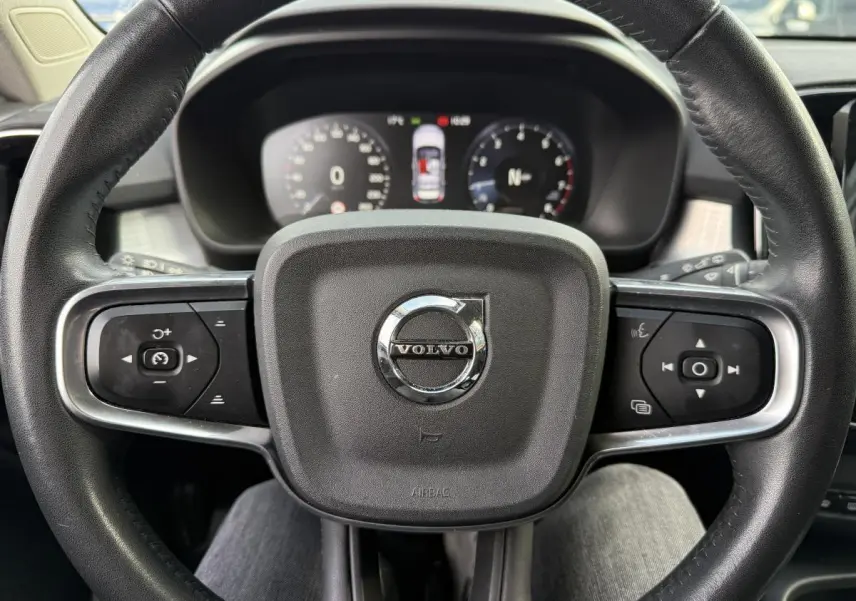 Volant cuir noir avec logo Volvo au centre, commandes intégrées, tableau de bord digital en arrière-plan.