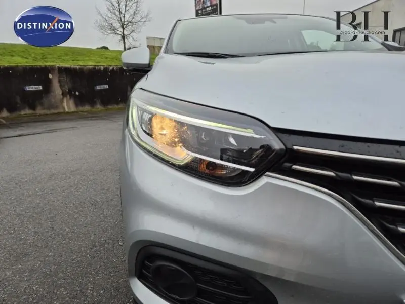 Gros plan sur le phare avant gauche allumé d’un Renault Kadjar blanc glacier, avec calandre et pare-chocs visibles.