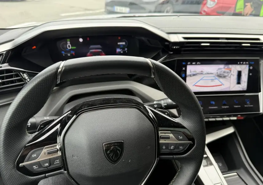 Vue rapprochée du volant cuir perforé noir et du tableau de bord numérique de la Peugeot 408 blanche 2024.