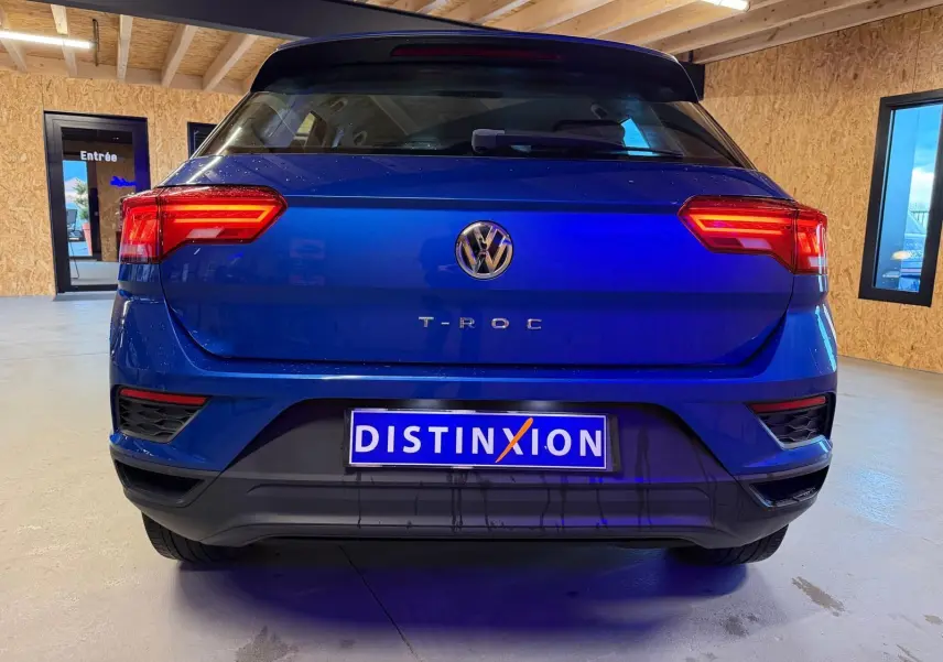 Vue arrière d'un Volkswagen T-Roc bleu 2019 avec feux arrière allumés et plaque Distinxion visible en intérieur.