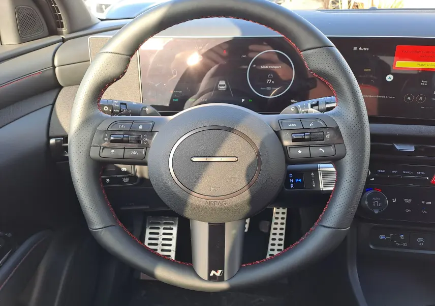 Vue frontale du volant noir perforé avec surpiqûres rouges du Hyundai Tucson 2025, tableau de bord numérique et commandes intégrées visibles.