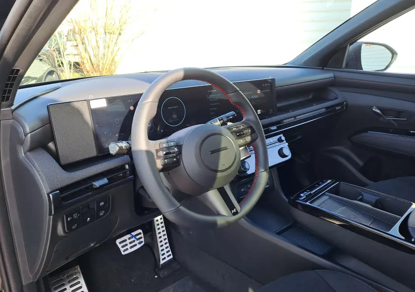 Vue intérieure côté conducteur du Hyundai Tucson 2025, tableau de bord noir avec écran digital large et volant cuir à surpiqûres rouges.