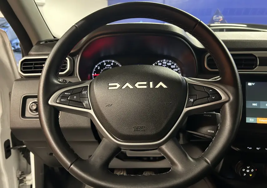 Vue rapprochée du volant noir du Dacia Duster 2023 blanc, avec commandes intégrées et tableau de bord en arrière-plan.