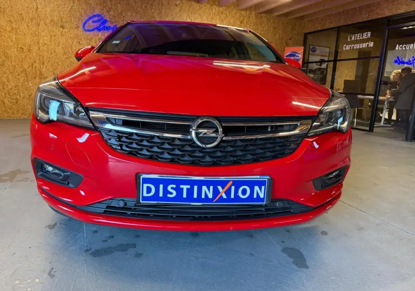Vue avant d’une Opel Astra rouge 2016 avec calandre noire et plaque d’immatriculation Distinxion en intérieur d’atelier.