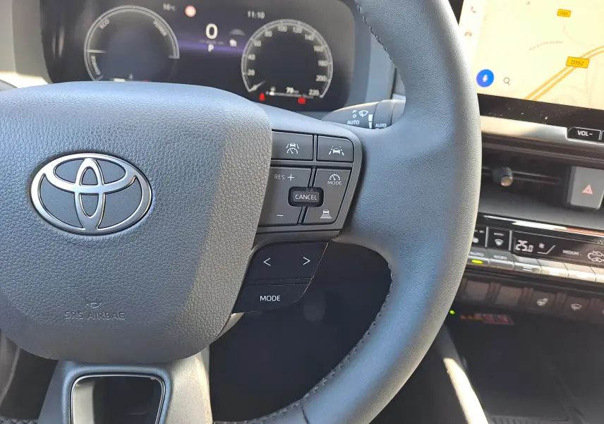 Gros plan sur le volant noir du Toyota C-HR gris minéral 2025, avec commandes intégrées et tableau de bord digital en arrière-plan.