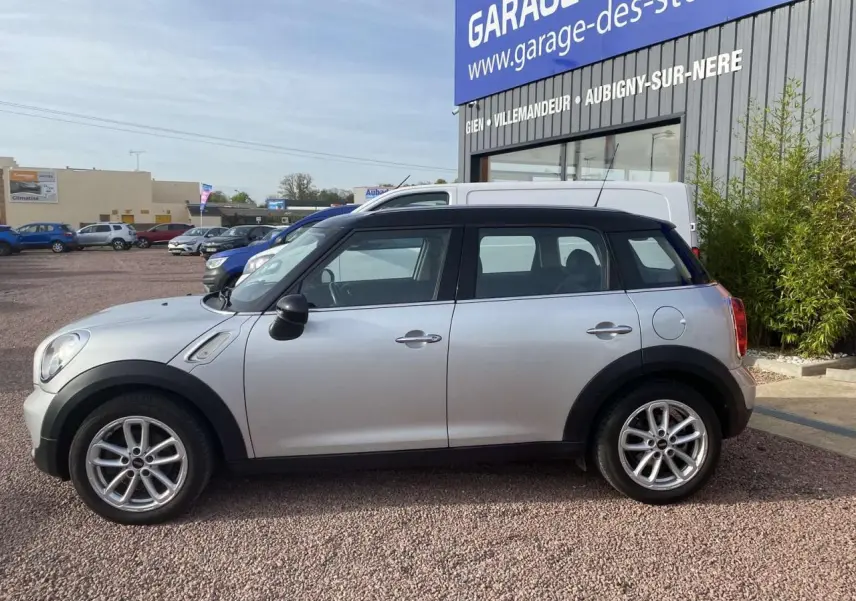 Profil côté gauche d'une MINI Countryman gris clair avec toit noir, garée devant un garage sur un sol gravillonné.