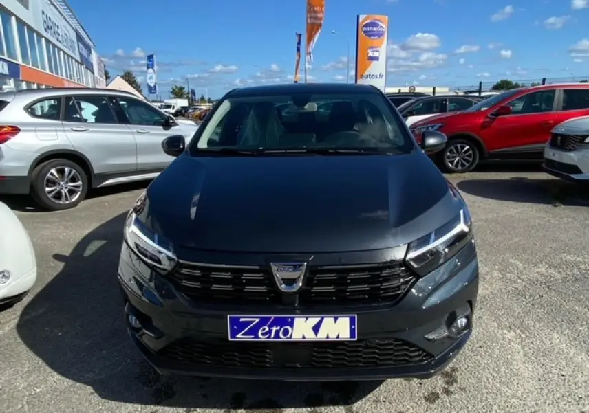 Vue de face d'une Dacia Logan gris foncé avec calandre noire et plaque "Zero KM" sur un parking extérieur.