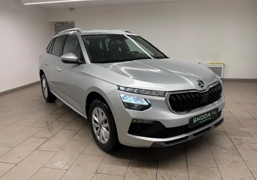 Vue 3/4 avant d'un Skoda Kamiq gris argent métallisé avec calandre noire et phares LED allumés, en intérieur.