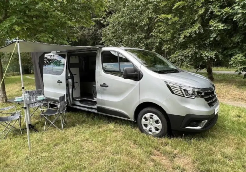 Renault Trafic Fourgon gris clair en 3/4 avant droit, porte latérale ouverte avec auvent et mobilier de camping installé.