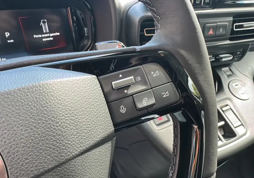 Gros plan sur le volant du Citroën Berlingo 2025 avec commandes intégrées et tableau de bord numérique affichant porte avant gauche ouverte.