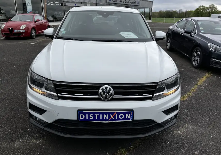 Volkswagen Tiguan blanc vu de face avec feux allumés, plaque Distinxion et parking de concession en arrière-plan.