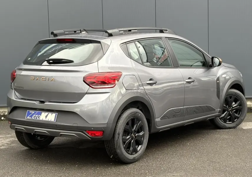 Vue 3/4 arrière droite d'une Dacia Sandero Stepway 2025 gris schiste avec jantes noires et barres de toit longitudinales.