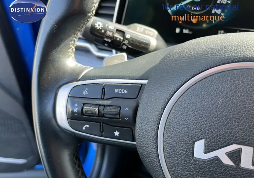 Gros plan sur le volant multifonction noir du Kia Sportage bleu fusion métal, avec commandes audio et tableau de bord en arrière-plan.