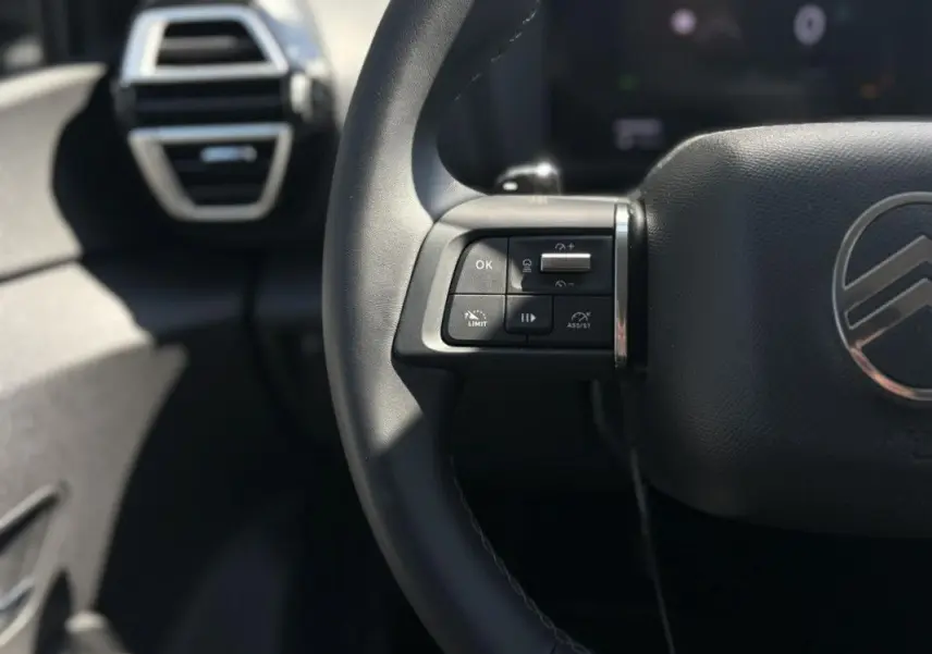 Gros plan sur la partie gauche du volant noir de la Citroën C4 Hybrid 145 ch 2025 avec commandes intégrées et logo centré.