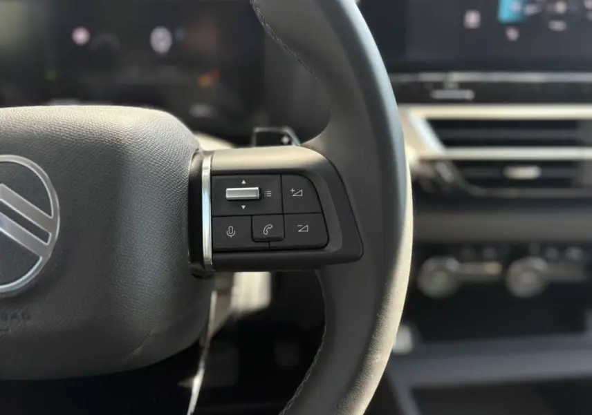 Gros plan sur les commandes gauche du volant noir de la Citroën C4 Hybrid 145 CH, avec tableau de bord flou en arrière-plan.
