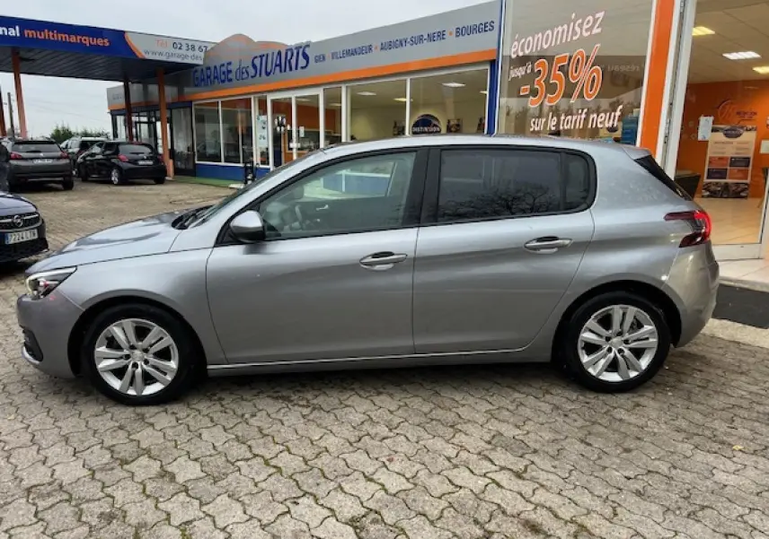 Peugeot 308 gris artense vue de profil côté gauche, garée devant un garage avec vitrine et panneau promotionnel.