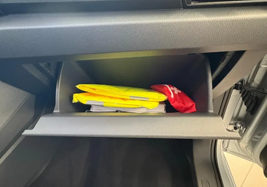 Compartiment de rangement ouvert côté passager du Volkswagen Caddy gris argent 2025, avec gilets de sécurité jaunes et trousse de secours rouge.