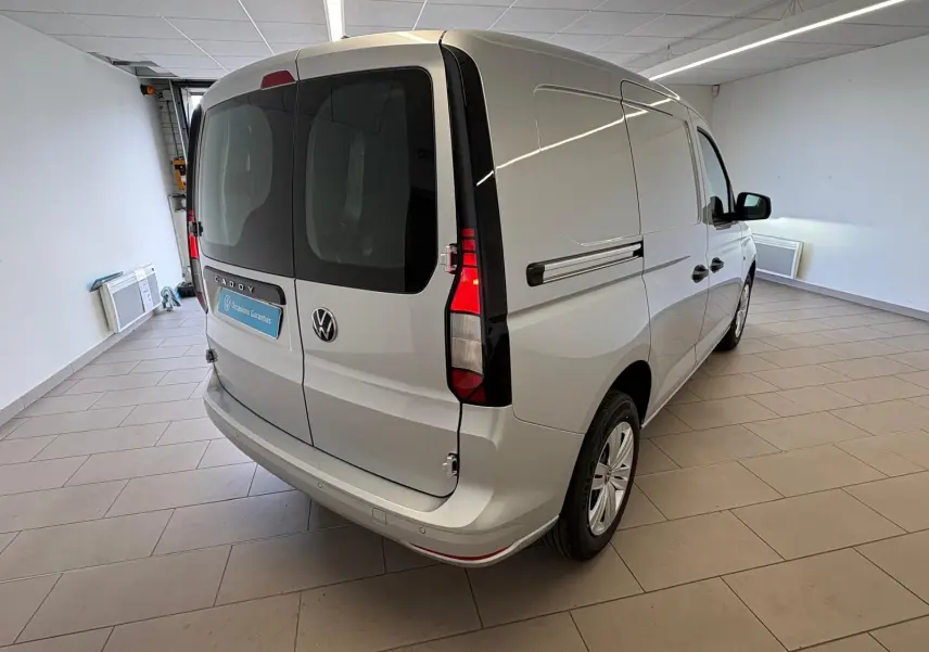 Vue 3/4 arrière droite d'un Volkswagen Caddy gris argent 2025 en version Business, avec portes arrière vitrées et feux verticaux.