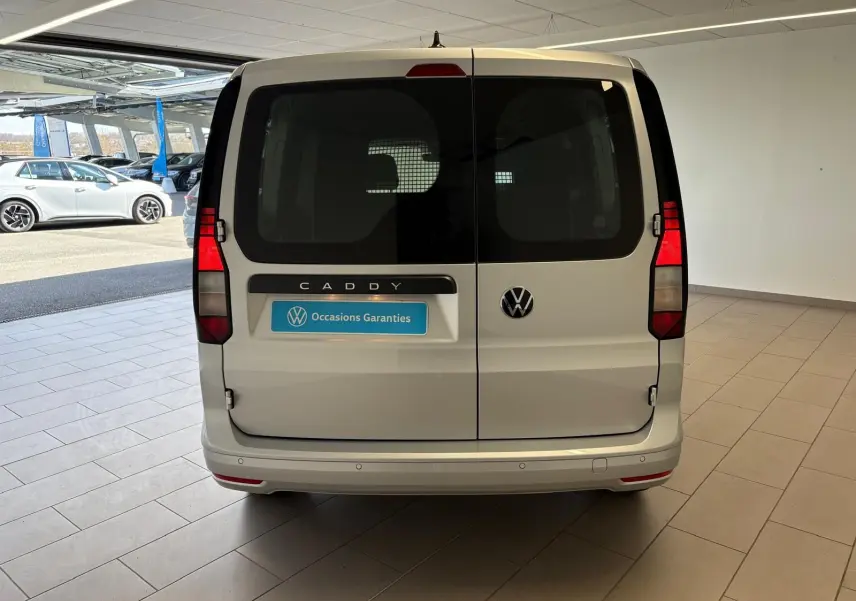 Vue arrière d'un Volkswagen Caddy gris argent 2025 avec portes battantes et logo VW visible en concession.