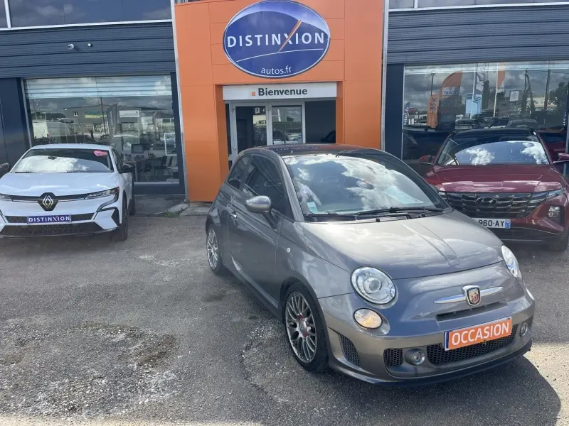 Vue 3/4 avant droite d'une ABARTH 500 gris foncé métallisé avec jantes alu et plaque "OCCASION" devant un garage.