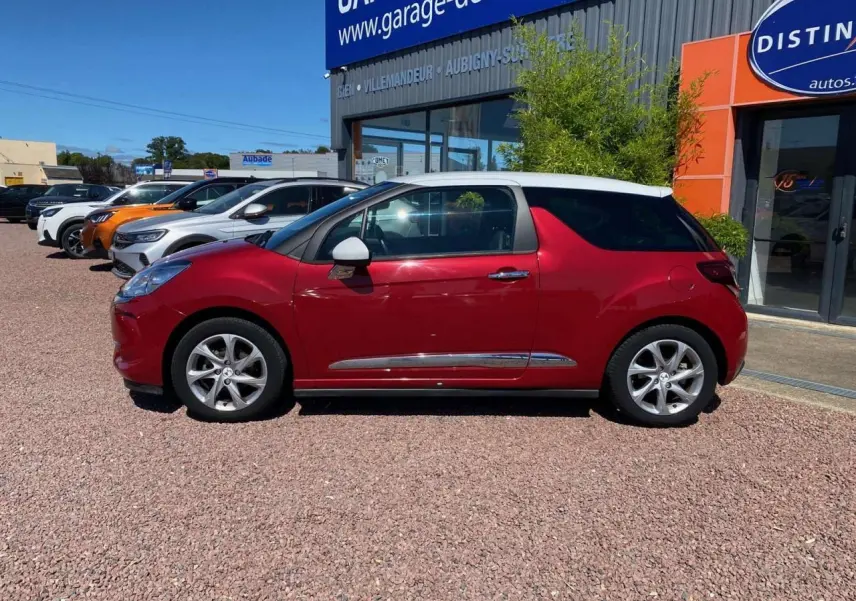 Profil côté gauche d'une DS3 rouge avec toit blanc, jantes alliage 16 pouces, stationnée devant un garage.