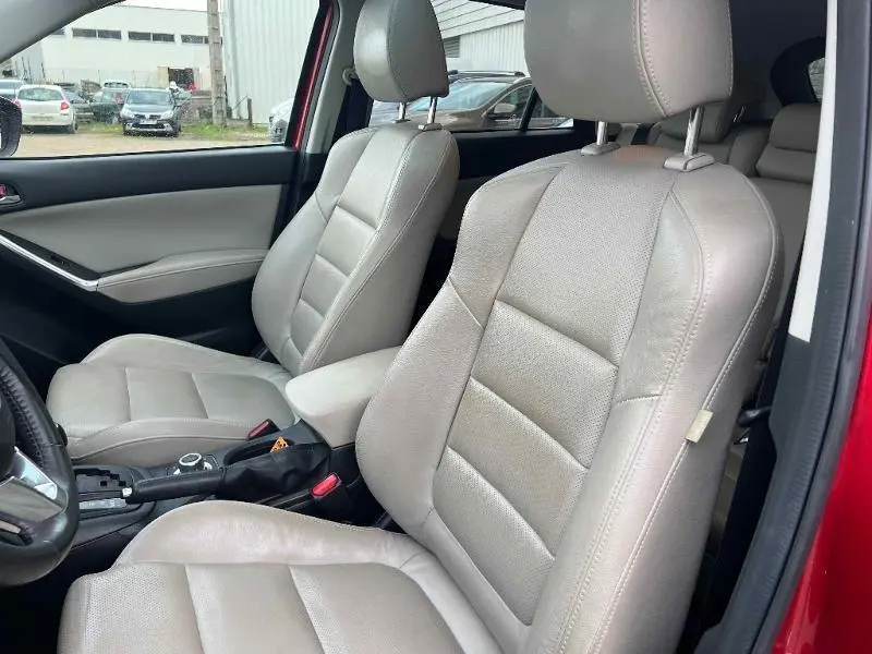 Vue intérieure côté conducteur du Mazda CX-5 rouge, mettant en valeur les sièges en cuir beige clair et l'accoudoir central.